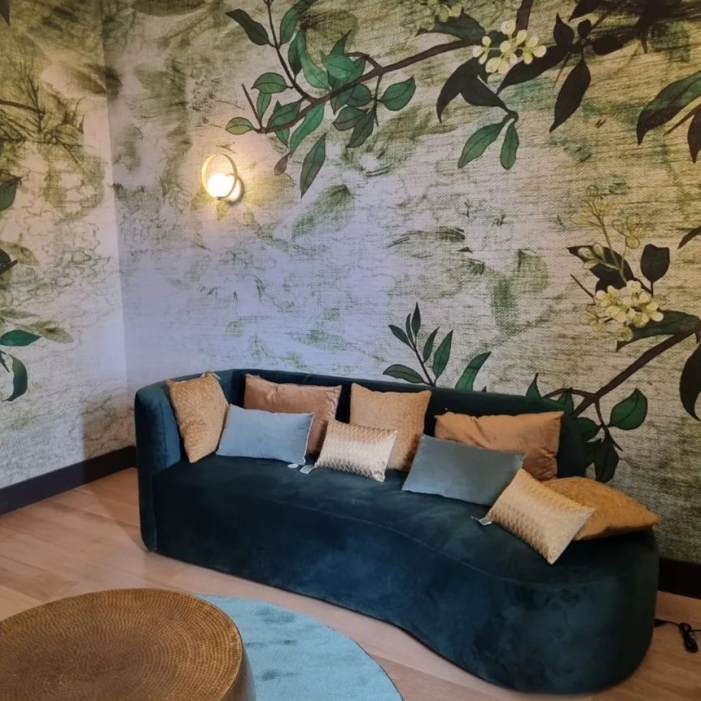Divano in velluto blu con cuscini dorati e parete decorata con motivi floreali.